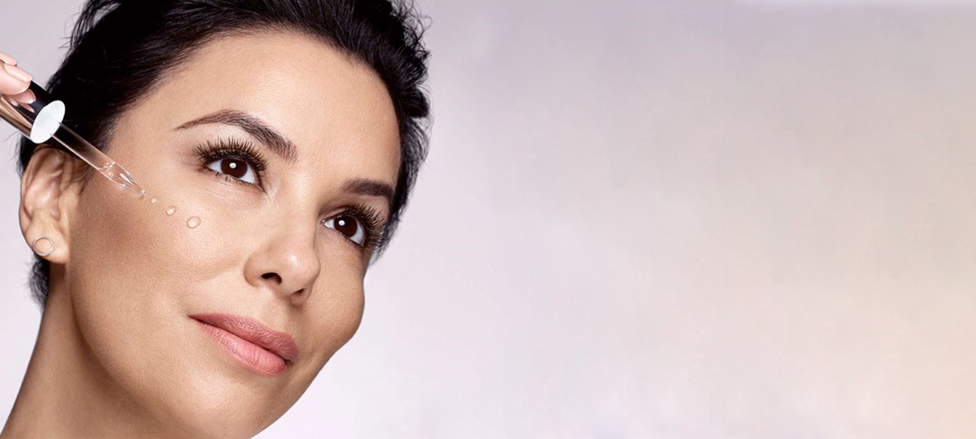 Dunkelhaarige Frau trägt sich Feuchtigkeitspflege mit Pipette auf die Wangen auf